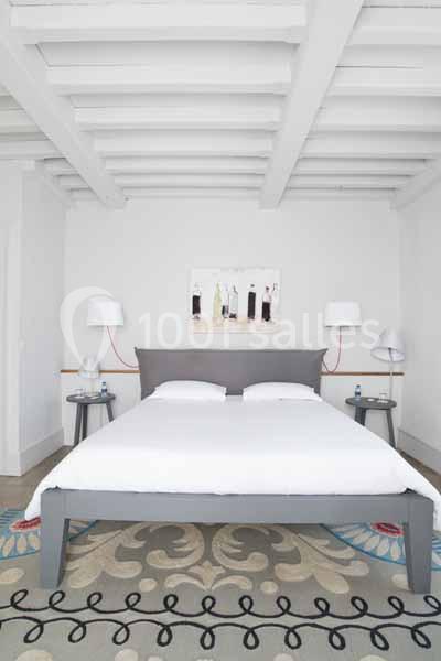 Lit double avec draps blancs, encadré par deux tables de chevet grises et des lampes, dans une chambre lumineuse au plafond…
