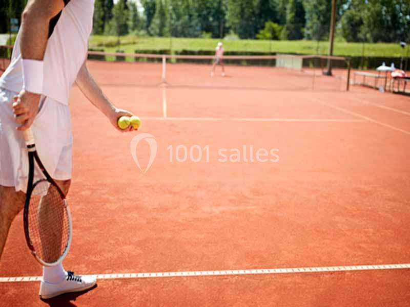 Un joueur de tennis en tenue blanche se prépare à servir sur un court en terre battue, avec un autre joueur en arrière-plan.