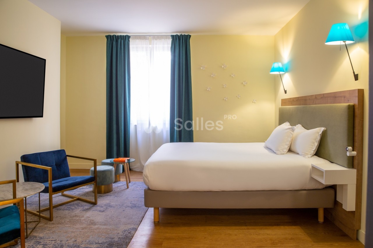 Chambre lumineuse avec lit double, fauteuils, table basse, rideaux bleus et appliques murales dans un décor moderne.