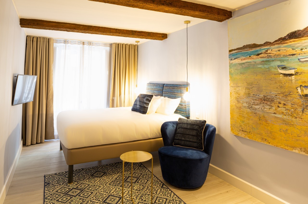 Chambre lumineuse avec lit double, fauteuil bleu, tableau coloré et grandes fenêtres avec rideaux beiges.