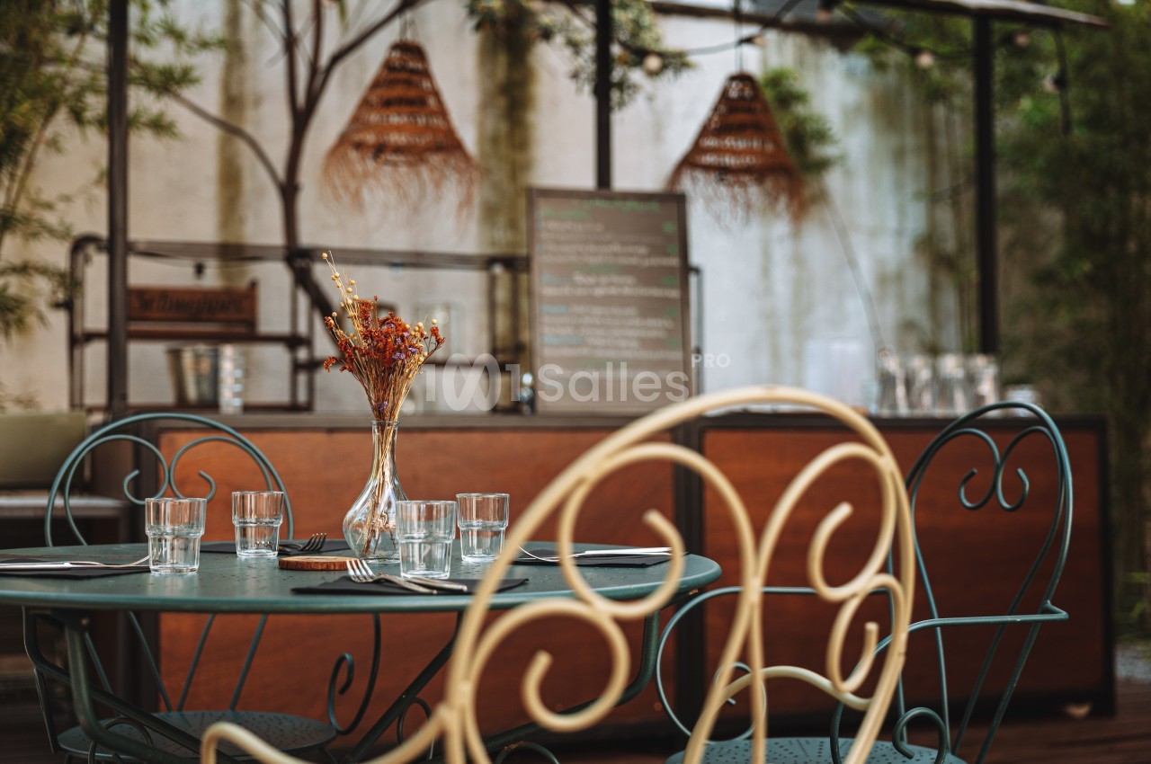 Table en métal avec verres et couverts, décorée d'un vase de fleurs séchées, dans une terrasse extérieure chaleureuse.