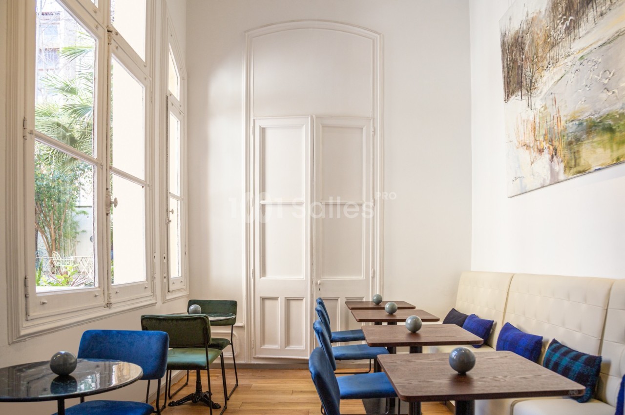 Salle lumineuse avec tables en bois, chaises colorées, banquette blanche et grandes fenêtres donnant sur un jardin.