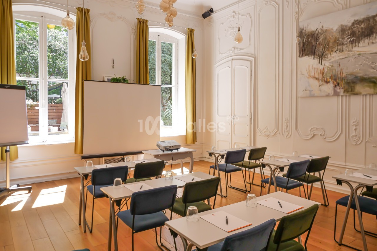 Salle de réunion lumineuse avec des chaises, des tables équipées de stylos et de verres, et un projecteur devant un écran.