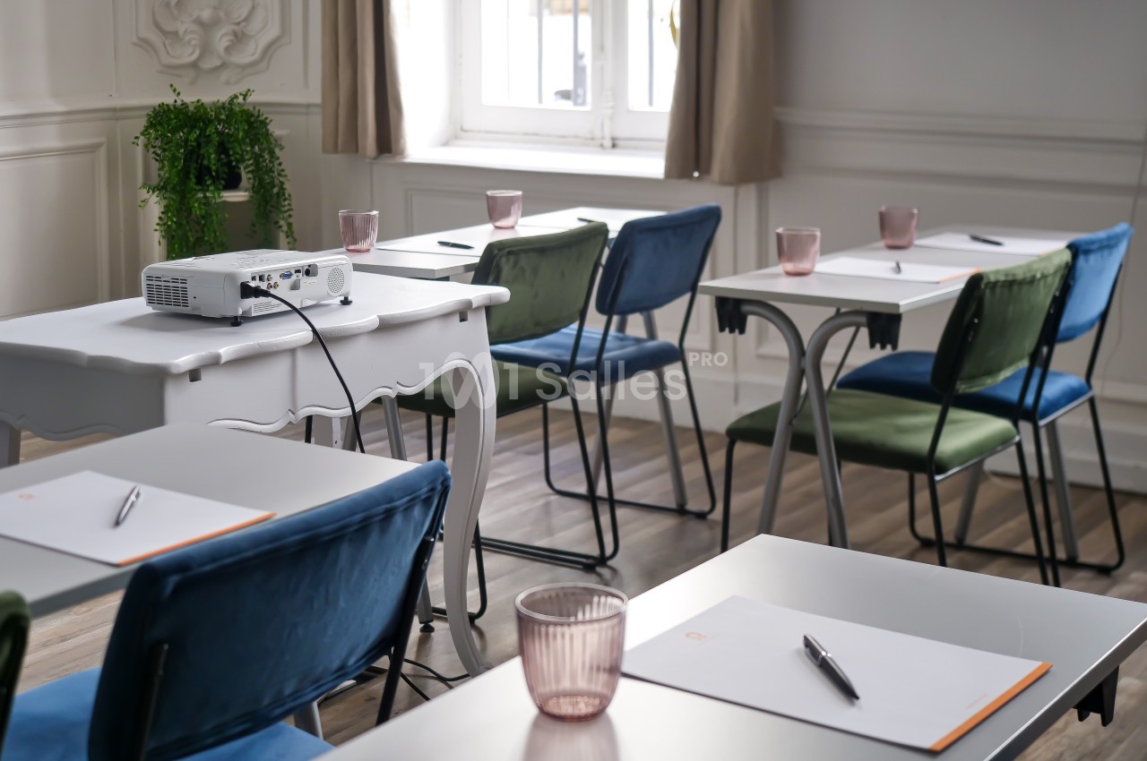 Salle de réunion lumineuse avec des chaises colorées, des tables équipées de blocs-notes, stylos et un projecteur.