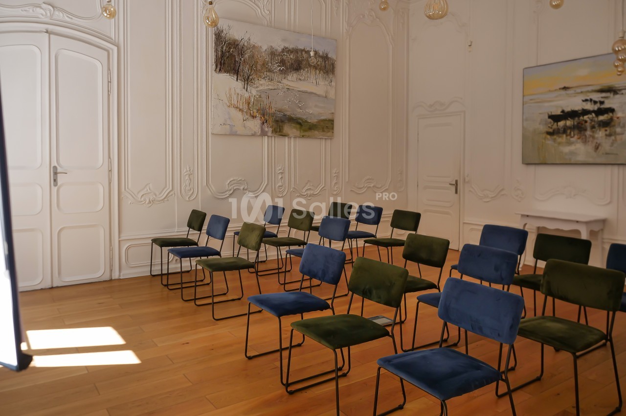 Salle lumineuse avec parquet, chaises alignées bleu et vert, murs ornés de moulures et tableaux paysagers.