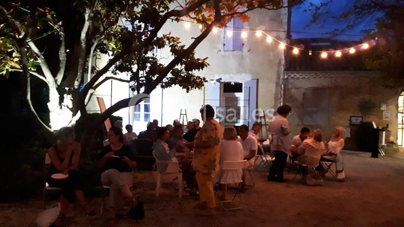 Groupe de personnes assises dans une cour éclairée par des guirlandes lumineuses, devant une maison en soirée.