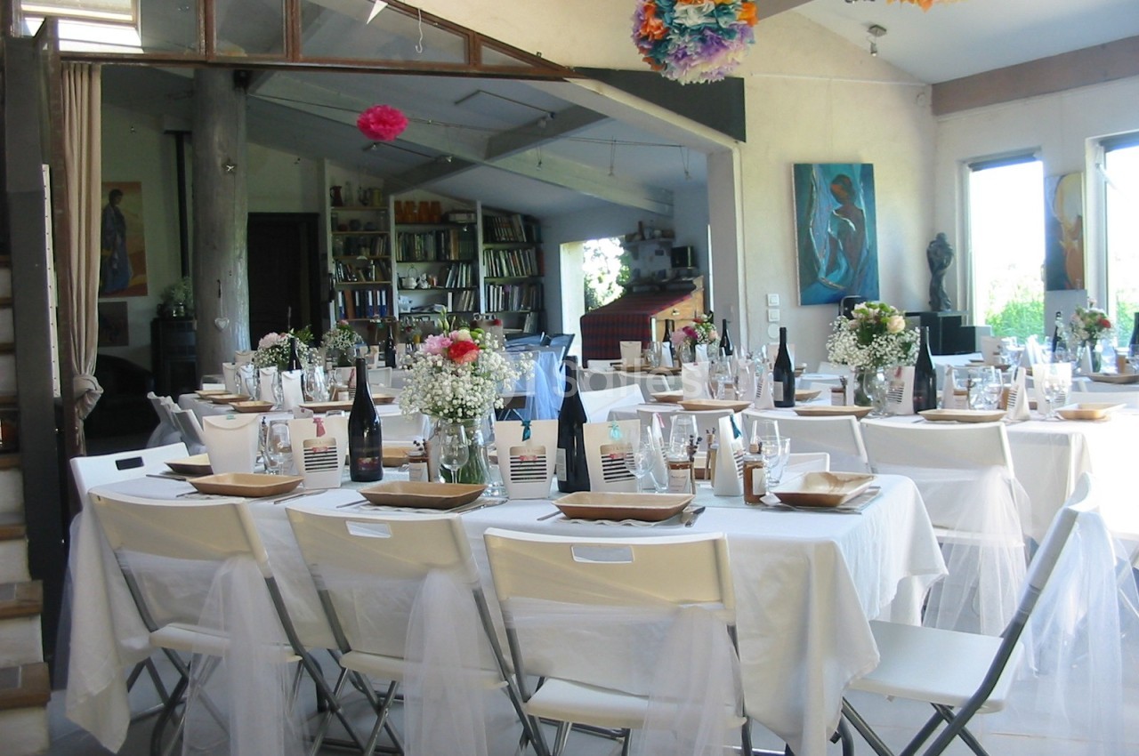 Salle décorée pour un repas festif avec tables dressées, nappes blanches, fleurs et chaises pliantes.