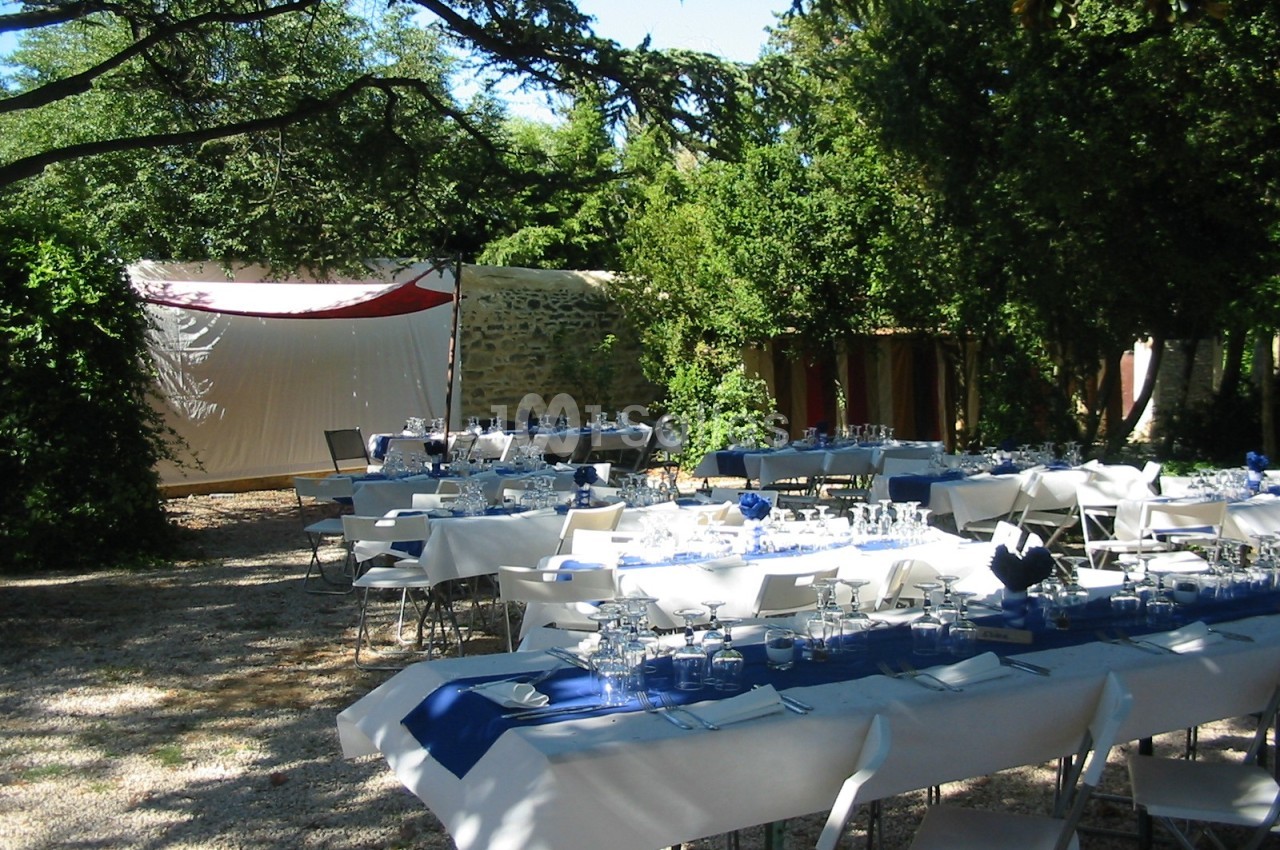 Tables dressées pour un repas en plein air sous des arbres, avec nappes blanches et décorations bleues.