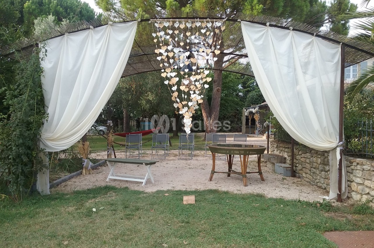 Structure en plein air avec rideaux blancs, décorations suspendues et mobilier de jardin sur sol en gravier.