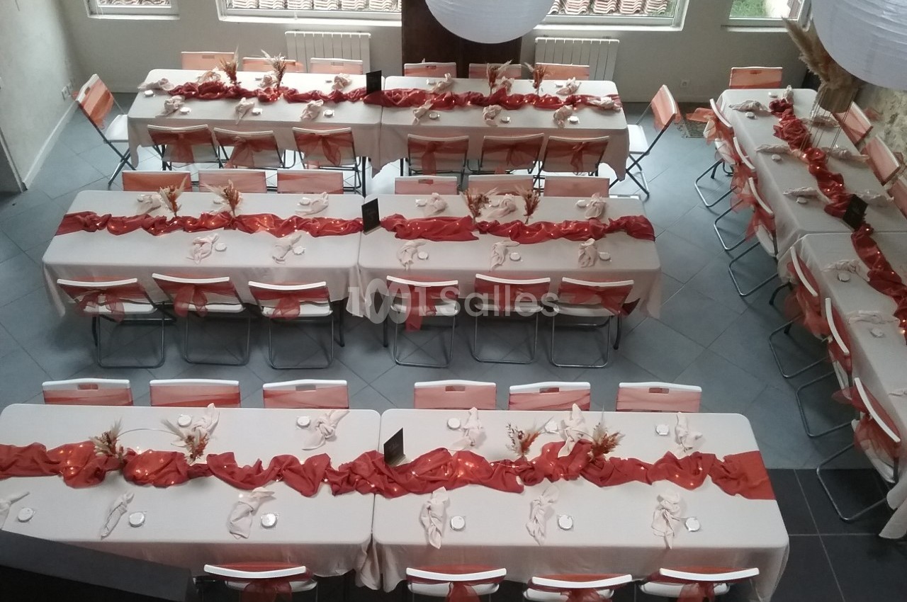 Salle décorée pour un événement, avec des tables alignées, nappes blanches et chemins de table rouges ornés de décorations.