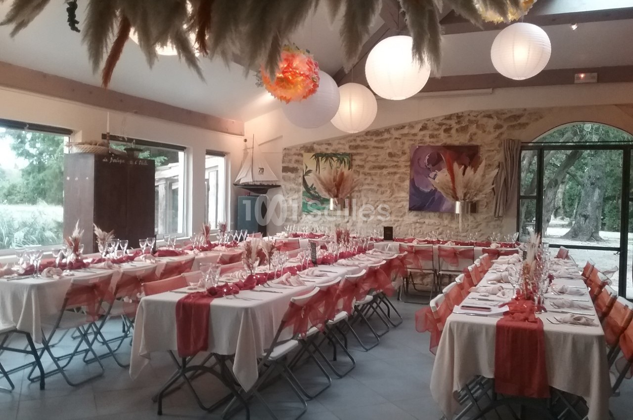 Salle décorée pour un événement, avec des tables alignées, nappes blanches, chemins de table rouges et lanternes suspendues.