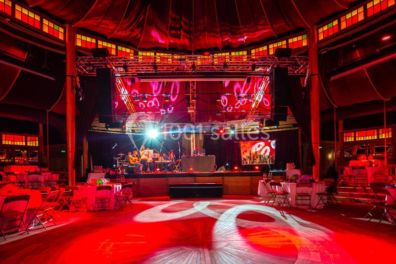 Salle de spectacle illuminée en rouge avec scène centrale, musiciens en arrière-plan et tables dressées autour.