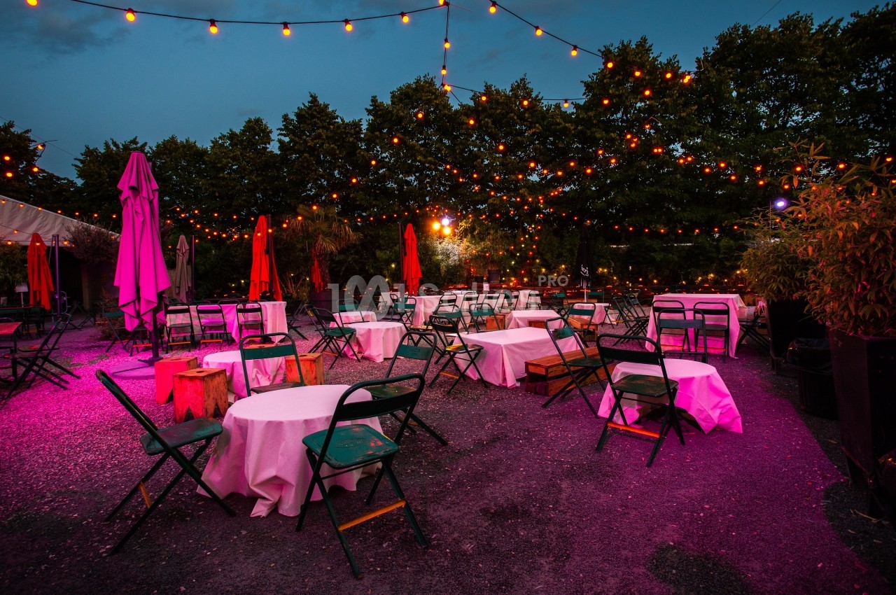 Terrasse extérieure avec tables rondes, chaises pliantes et guirlandes lumineuses suspendues, ambiance en soirée.