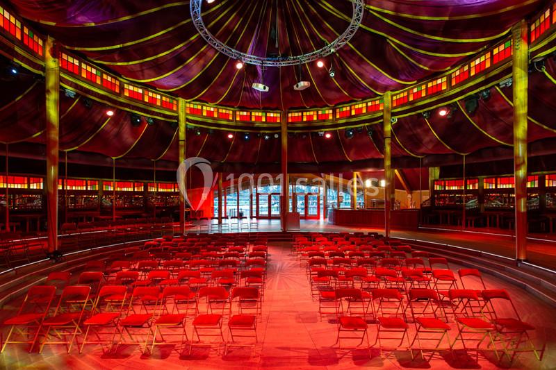 Intérieur d’un chapiteau avec des rangées de chaises rouges disposées face à une scène sous un éclairage coloré.