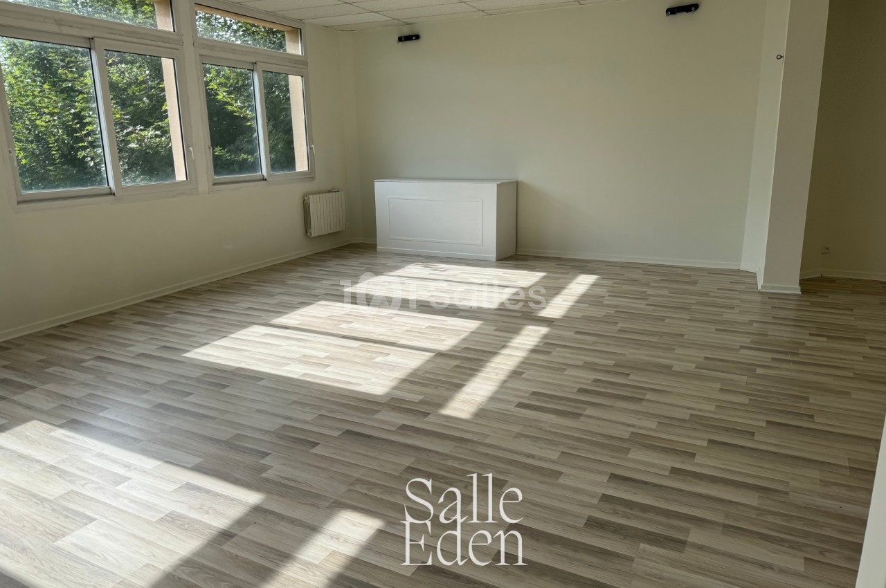 Salle vide avec parquet clair, murs blancs, grandes fenêtres laissant entrer la lumière naturelle et vue sur des arbres.