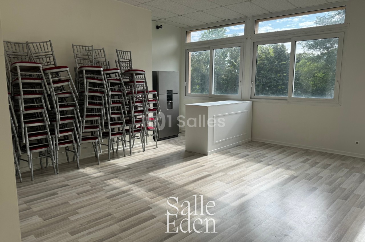 Salle lumineuse avec parquet clair, chaises empilées sur la gauche, comptoir blanc et grandes fenêtres donnant sur des…