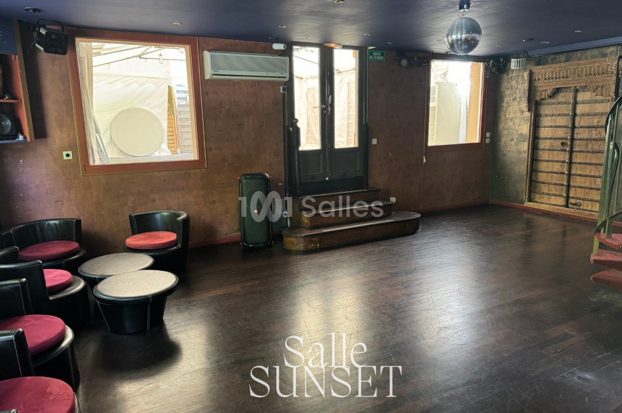 Salle vide avec parquet sombre, murs en bois, fauteuils noirs, tables basses, et une boule disco au plafond.