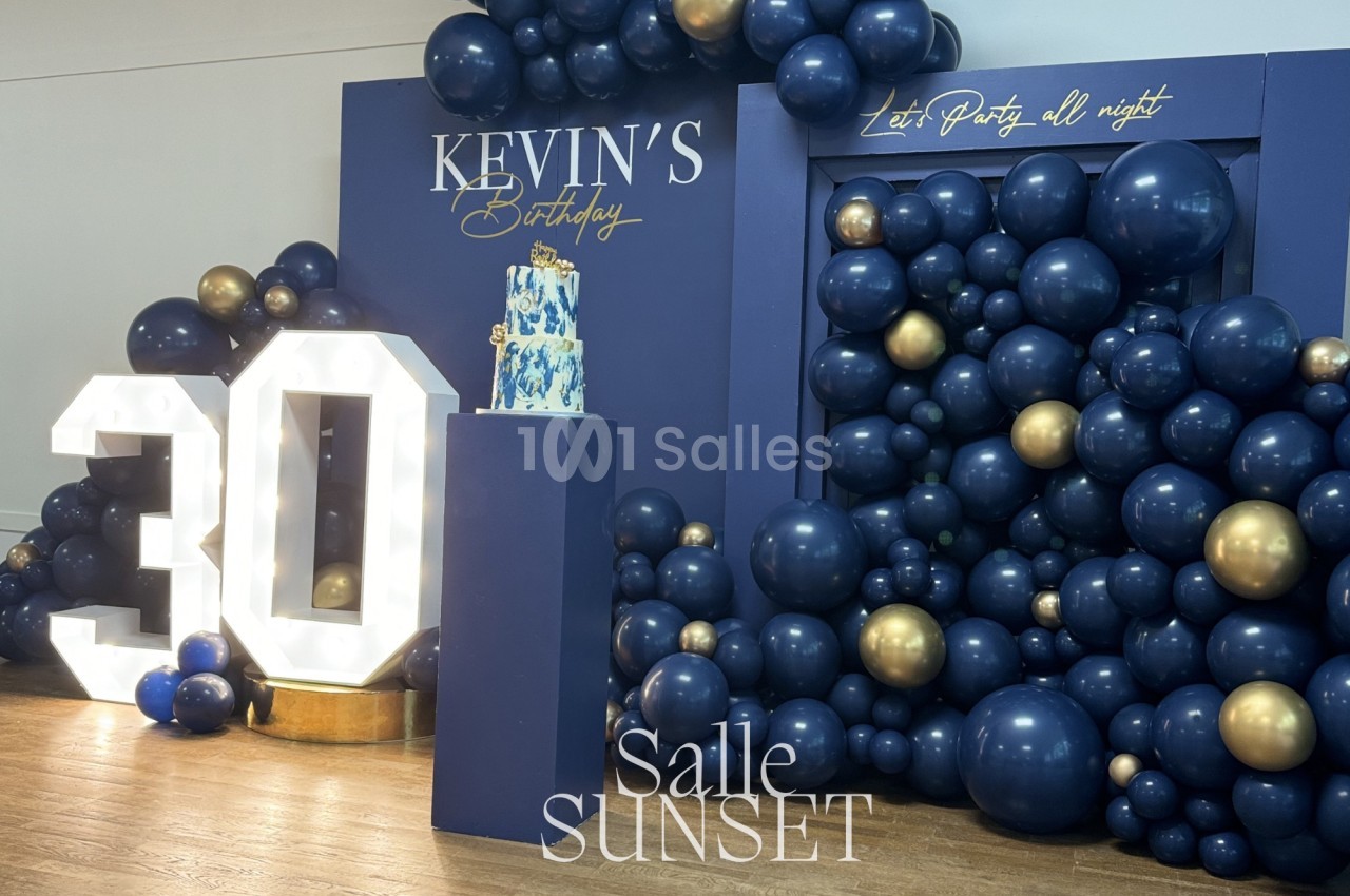 Décoration d'anniversaire avec ballons bleu marine et dorés, chiffre lumineux ’30’ et gâteau sur un présentoir.