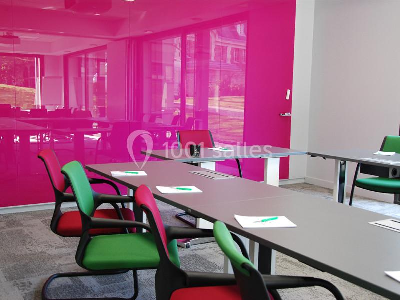 Salle de réunion moderne avec murs roses, tables grises, chaises colorées et documents posés sur les tables.