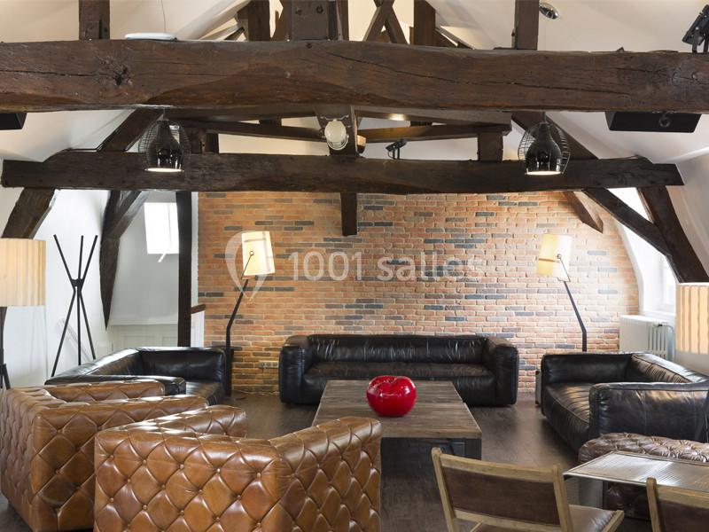 Salon avec poutres apparentes, murs en briques, fauteuils en cuir et table basse en bois, éclairé par des lampes modernes.
