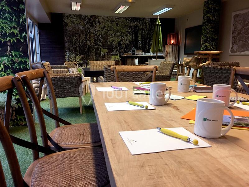 Salle de réunion avec table en bois, chaises en osier, mugs, carnets et décor mural représentant une forêt.