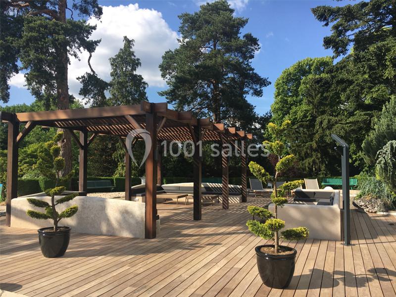 Terrasse en bois avec pergola en bois sombre, mobilier de jardin et plantes en pots, entourée de végétation dense.