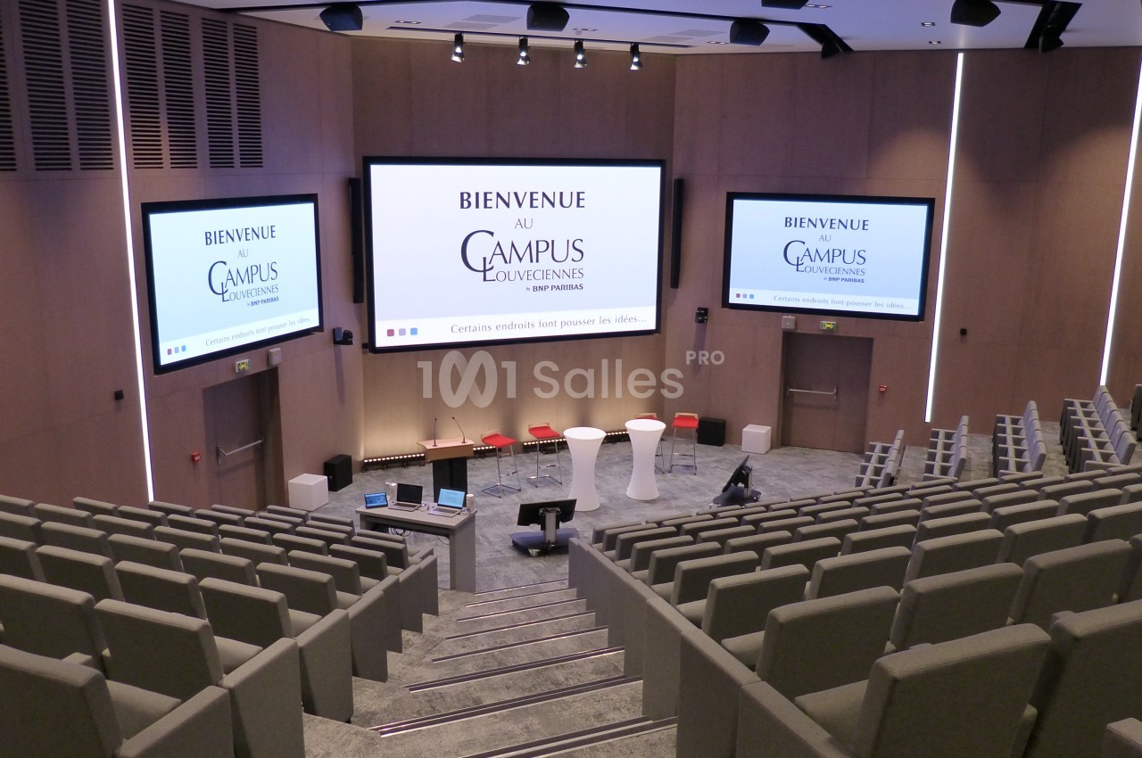Salle de conférence moderne avec sièges gris, écrans géants affichant ’Bienvenue au Campus Louvain’.