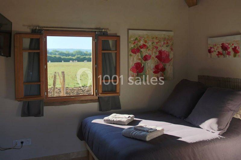 Vue d'une chambre lumineuse avec un lit double, des tableaux floraux et une fenêtre ouverte sur un paysage champêtre.