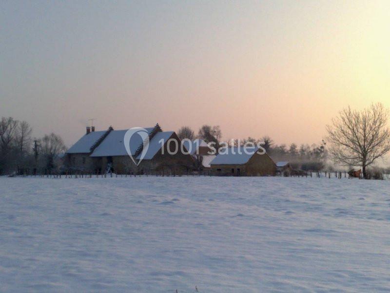 Ferme en pierre entourée de champs enneigés au lever du soleil, avec des arbres nus en arrière-plan.