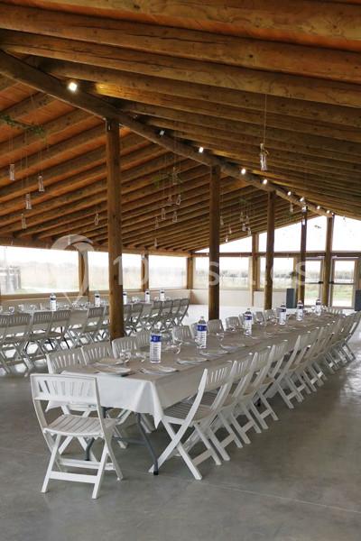 Salle avec charpente en bois, tables rectangulaires dressées avec nappes blanches et chaises pliantes blanches.