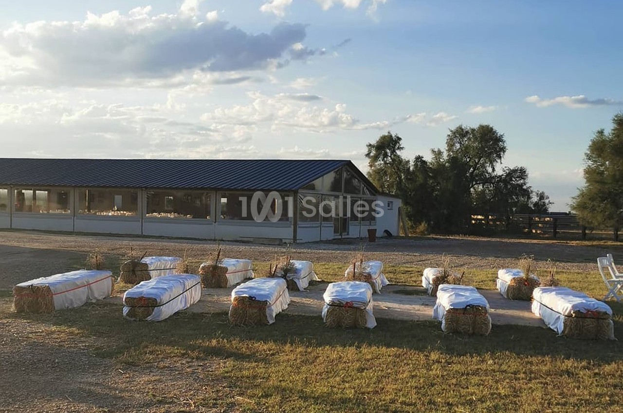 Bottes de paille recouvertes de tissu blanc disposées en rangées devant un bâtiment avec toit en tôle, sous un ciel dégagé.