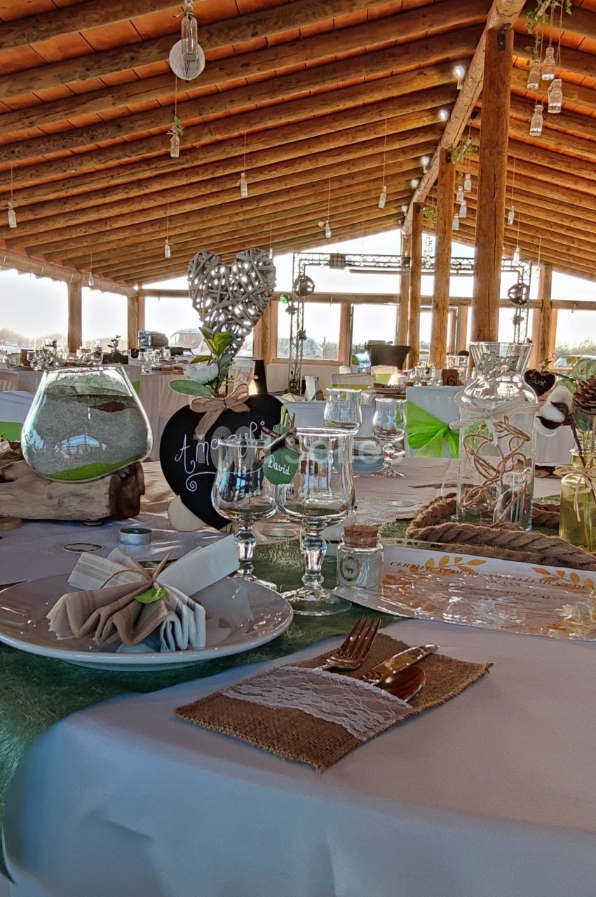 Table décorée pour un événement avec nappes blanches, couverts dorés, verres, et ornements sous un toit en bois.