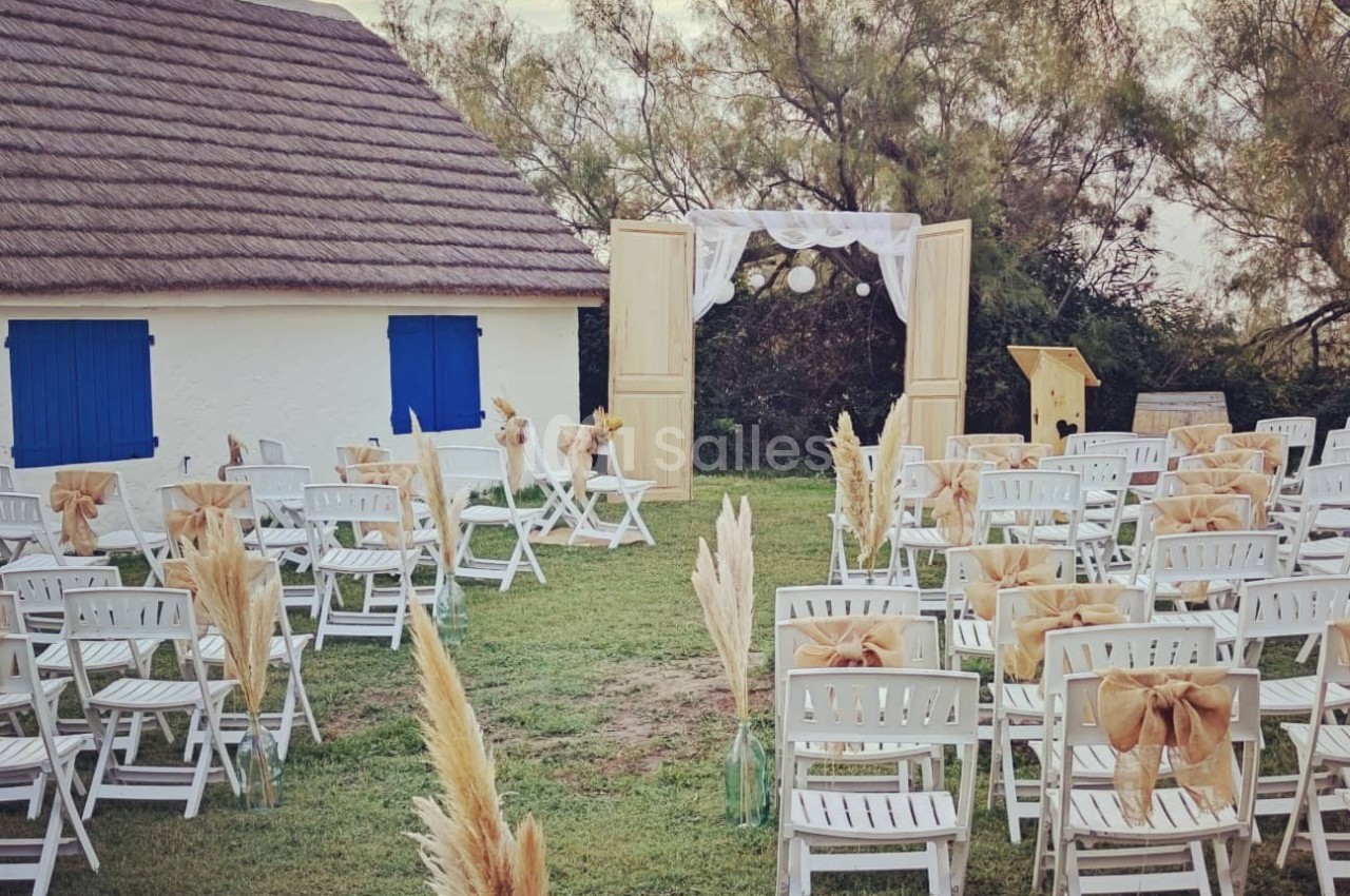 Chaises blanches décorées disposées en extérieur devant une arche ornée de tissu, près d'une maison au toit de chaume.