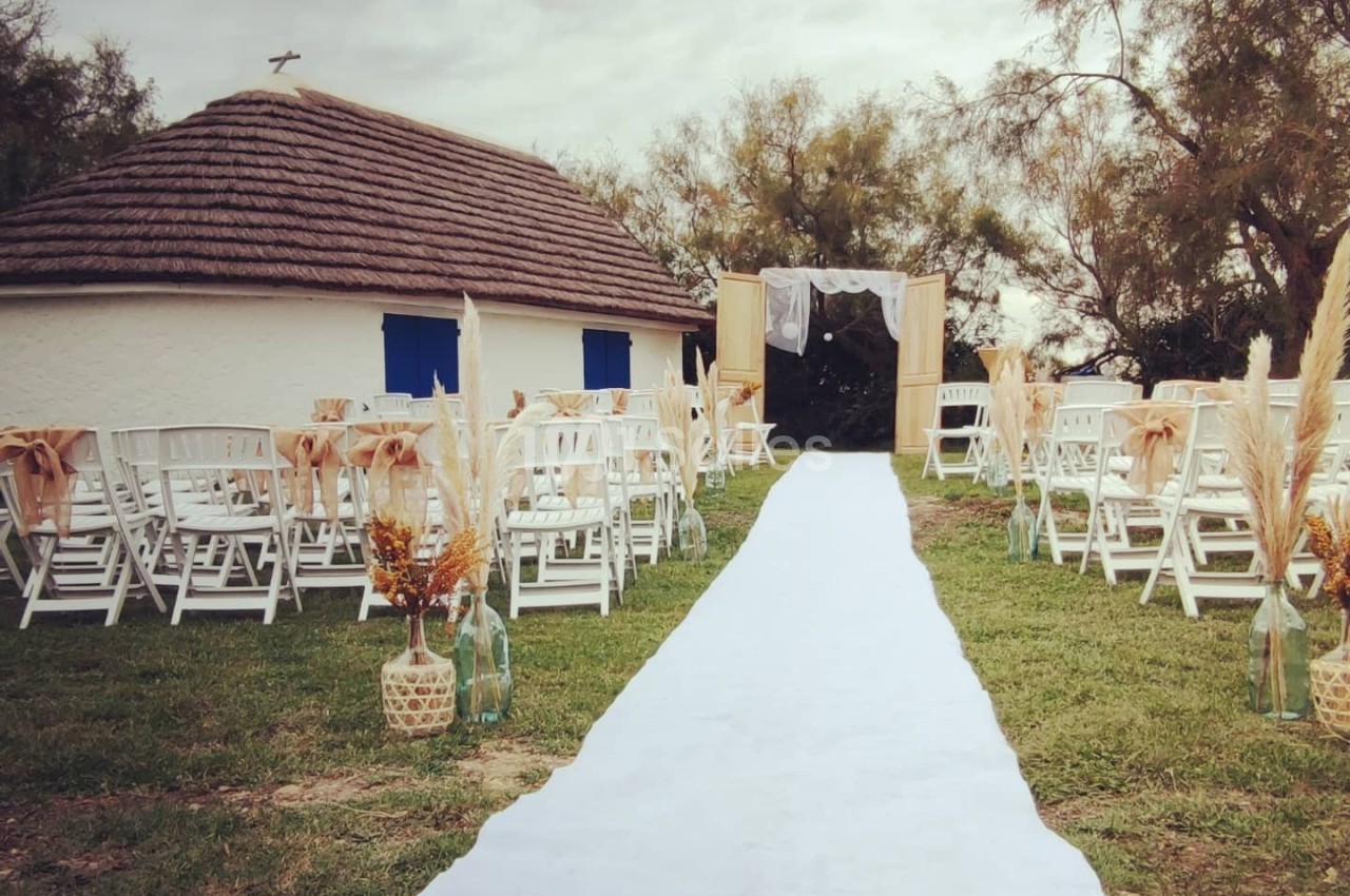 Allée blanche menant à une arche décorée, entourée de chaises blanches, devant une petite maison au toit de chaume.