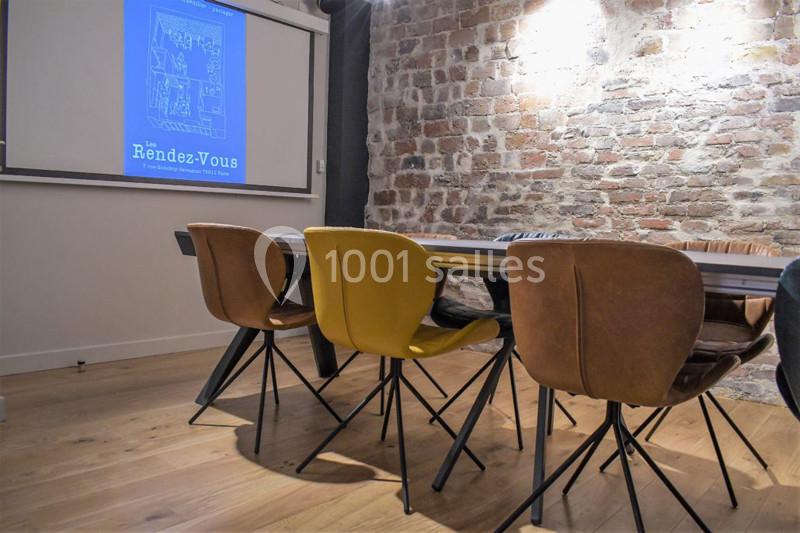 Salle de réunion avec table en bois, chaises modernes et mur en briques, écran de projection allumé.