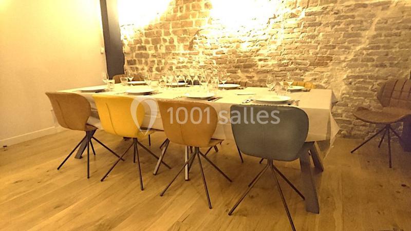 Table dressée avec vaisselle et verres, entourée de chaises colorées dans une pièce avec mur en briques apparentes.