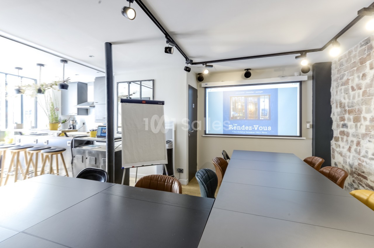 Salle de réunion moderne avec table noire, chaises, écran de projection, paperboard et cuisine ouverte en arrière-plan.