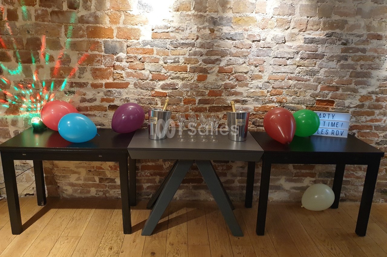 Tables noires décorées de ballons colorés, seaux à champagne et verres, devant un mur de briques.