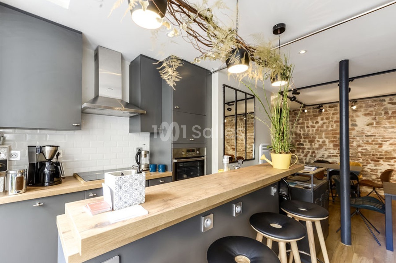 Cuisine moderne avec comptoir en bois, tabourets noirs, murs en briques apparentes et éclairage suspendu.