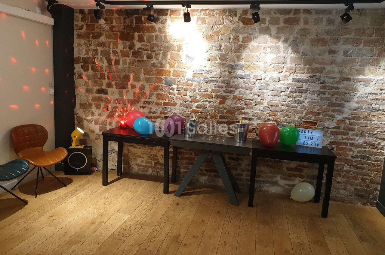 Salle décorée pour une fête avec des ballons colorés sur une table devant un mur en briques.