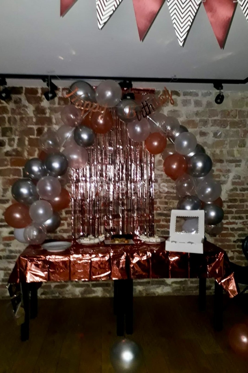Décoration d'anniversaire avec ballons argentés et cuivrés, guirlande et table dressée devant un mur de briques.