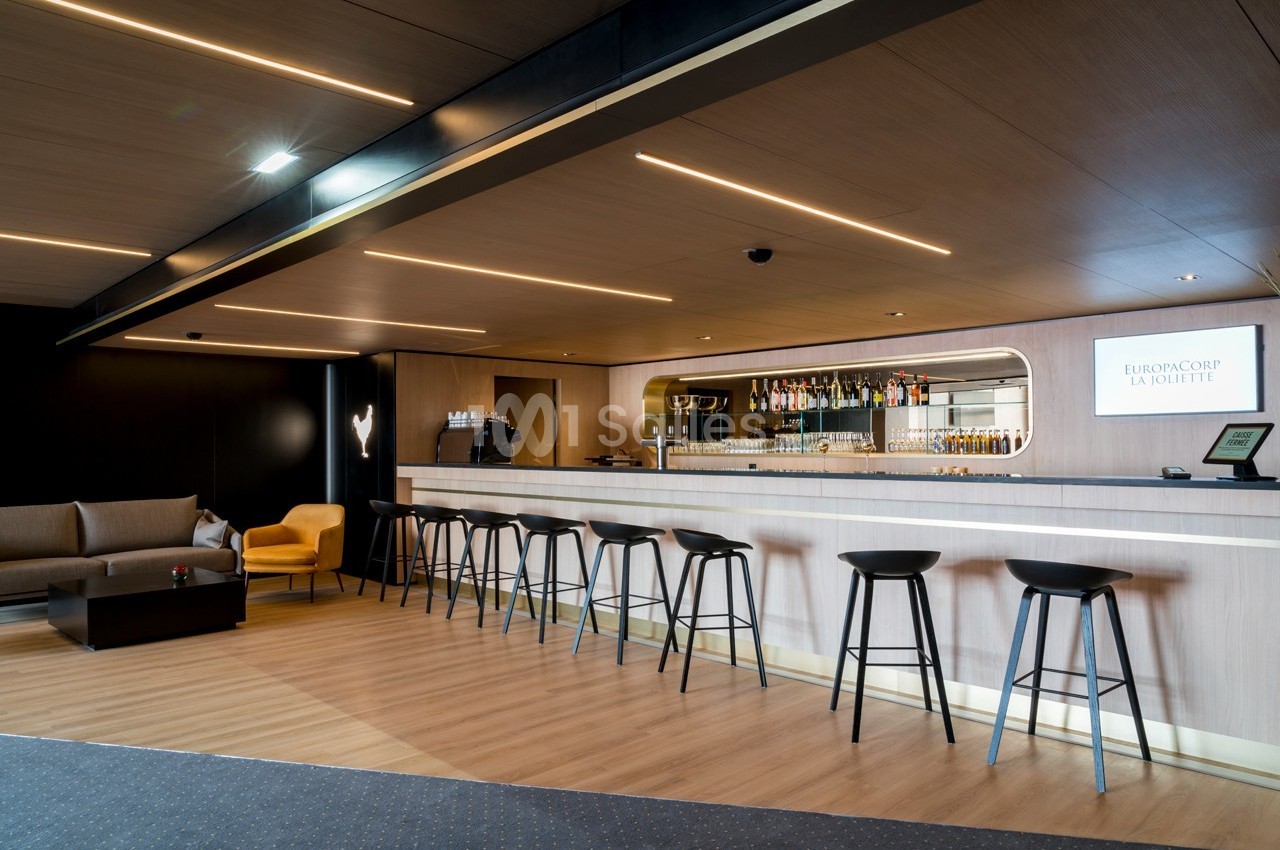 Bar moderne avec comptoir en bois clair, tabourets noirs, étagères de bouteilles et coin salon avec fauteuils.