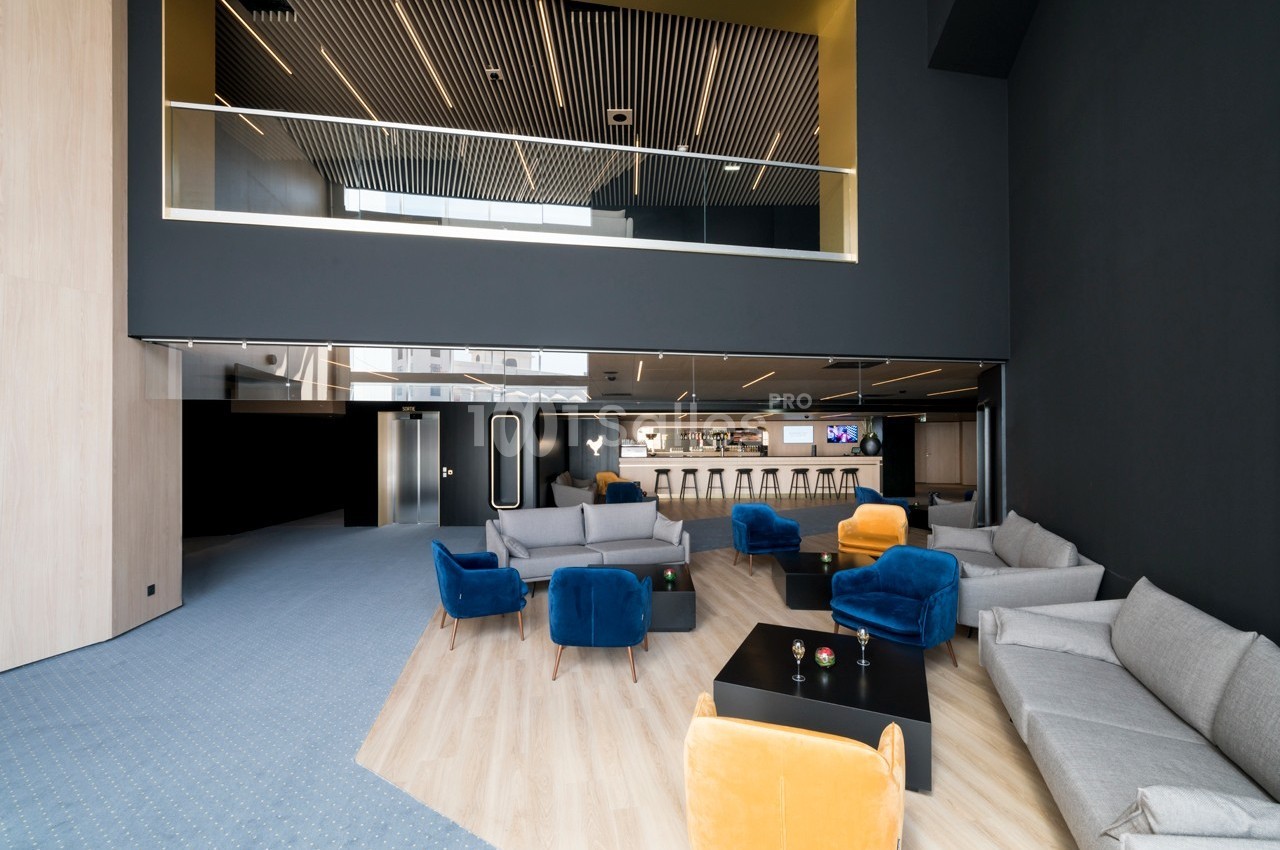 Espace lounge moderne avec fauteuils colorés, tables basses et bar en arrière-plan sous un plafond à lames en bois.