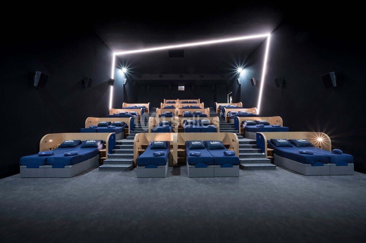 Salle de cinéma avec des lits bleus alignés en gradins, éclairage tamisé et projecteurs muraux.