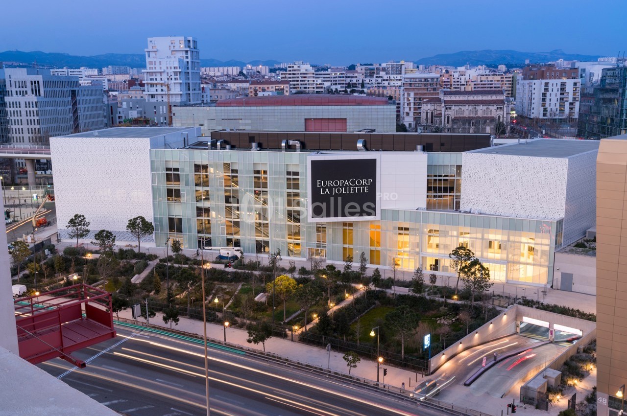 Façade du cinéma EuropaCorp La Joliette, entourée de bâtiments modernes et d'une route éclairée au crépuscule.