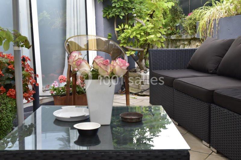 Salon de jardin avec table en verre, vase de roses, assiettes et canapé, entouré de plantes et fleurs.