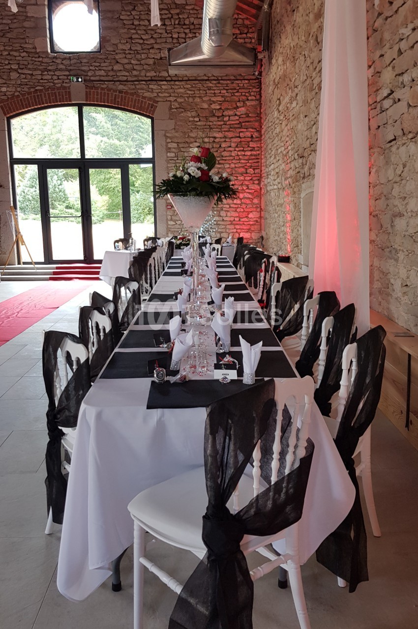 Salle de réception avec une longue table dressée, nappes blanches, chaises décorées de nœuds noirs et centre de table floral.