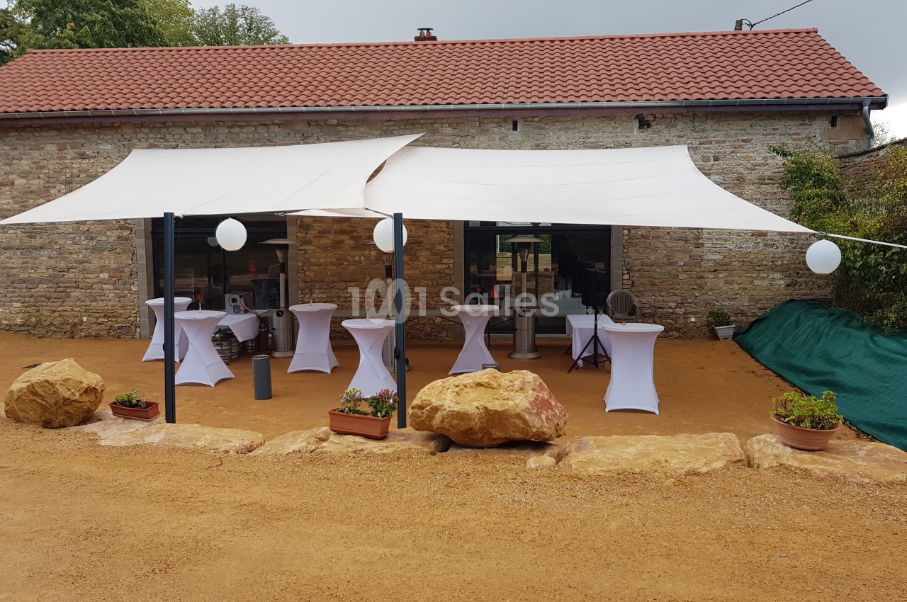 Terrasse aménagée avec des voiles d'ombrage, tables hautes habillées de housses blanches et décorations extérieures.