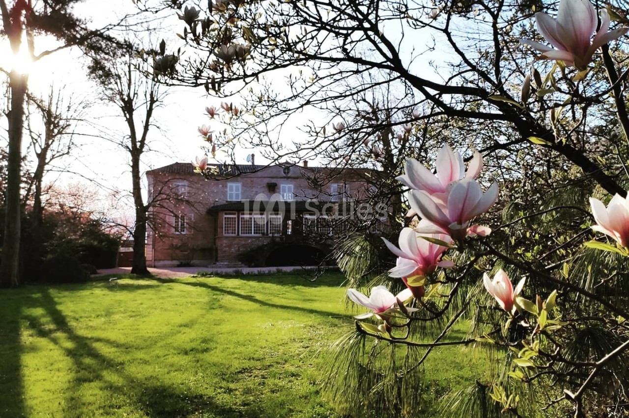 Maison en pierre entourée d'un jardin verdoyant, avec des magnolias en fleurs au premier plan et un soleil couchant.