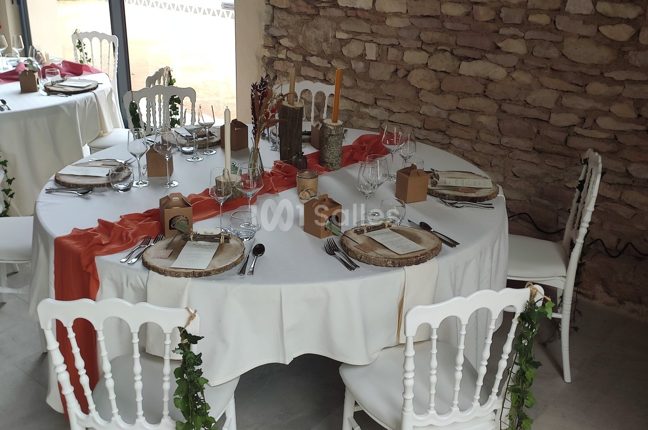 Salle décorée pour un repas festif avec tables rondes dressées, nappes blanches, chemins de table orange et chaises blanches.
